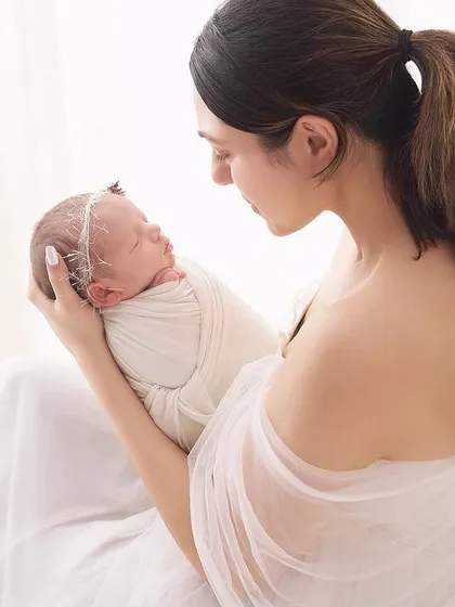 A quiet moment between mother and newborn, captured from the side. The soft white fabrics and natural light from the window create a peaceful and angelic atmosphere for this newborn session.