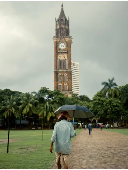 A person walks with an umbrella on a path in Oval Maidan, with the Rajabai Clock Tower in the background. This image beautifully juxtaposes a slice of everyday life with one of Mumbai's iconic landmarks.