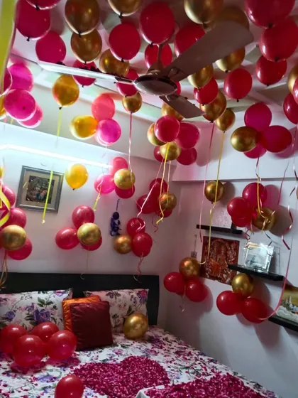 A vibrant birthday room filled with red, pink, and gold balloons. The bed is covered with rose petals, making it a colorful and romantic surprise.