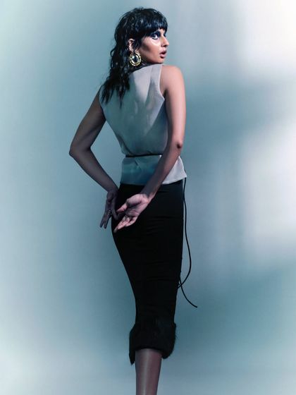 An editorial shot with a moody, blue-toned lighting setup. The styling combines a sheer, sleeveless top with a black pencil skirt, creating a look that is both elegant and mysterious.