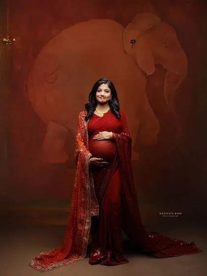A standing portrait of the mother-to-be in a red saree, her hands gently holding her baby bump. The elephant motif backdrop adds a classic Indian touch.