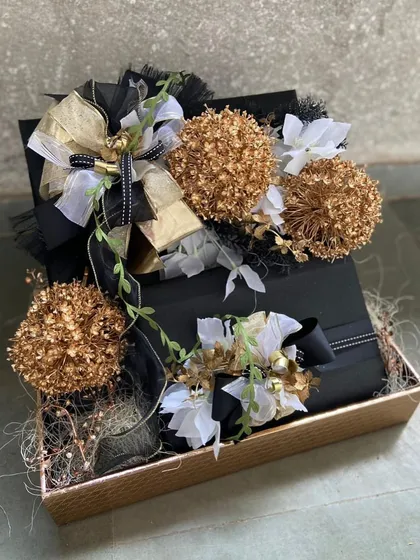 A dramatic and elegant black and gold themed hamper. This arrangement features striking golden pom-poms and delicate white flowers against a black gift box, making it a perfect choice for a sophisticated evening event or a glamorous celebration.