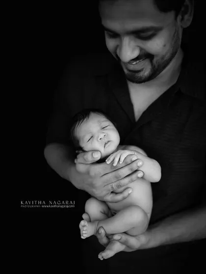 A black and white portrait of a father holding his nine-day-old newborn. The dramatic lighting emphasizes the protective and loving nature of his embrace.