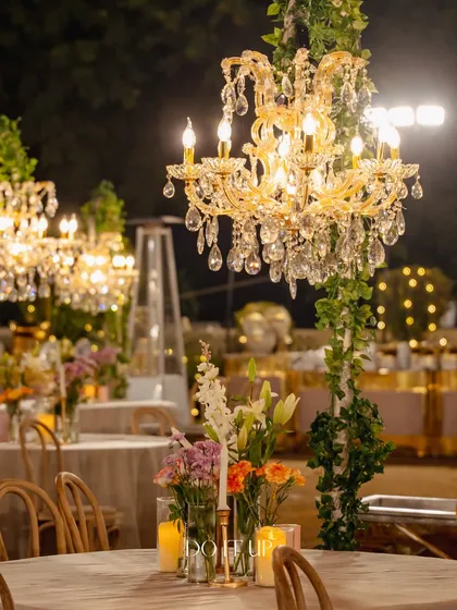 An elegant table setting at the outdoor Jaipur celebration. A crystal chandelier on a vine-wrapped stand serves as a unique and glamorous centerpiece, illuminating the table with a warm glow.