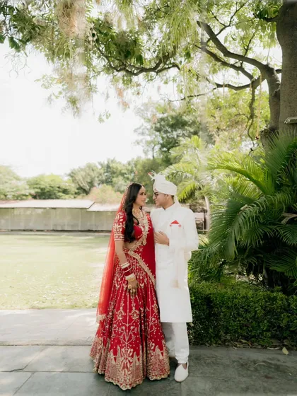 A classic portrait of the couple under a tree, their traditional red and white outfits standing out against the lush greenery.