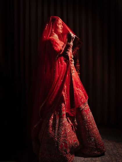 A dramatic bridal portrait with the bride standing in near darkness, her form illuminated by a single light source. The red of her lehenga and the silhouette of her veil create a powerful and artistic image.