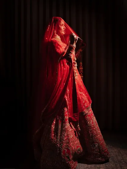 A dramatic bridal portrait with the bride standing in near darkness, her form illuminated by a single light source. The red of her lehenga and the silhouette of her veil create a powerful and artistic image.