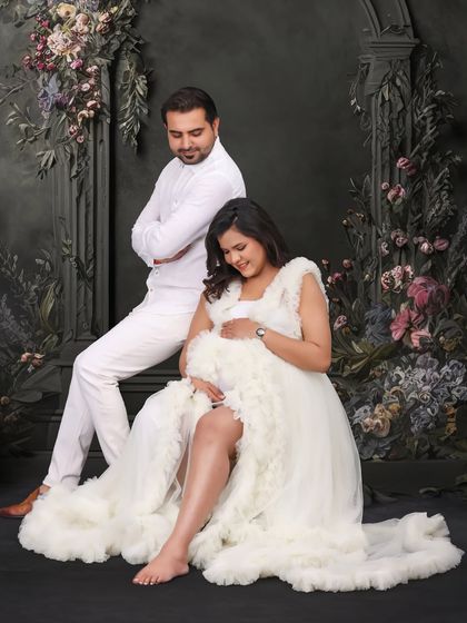 A relaxed and happy pose from a studio maternity shoot. The couple is dressed in all white, seated before a dark floral archway, creating a striking and artistic portrait.