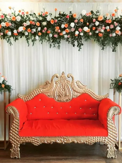An elegant and simple stage design for an engagement or reception. The backdrop of white drapes is accented with a beautiful floral arch, and a classic red and gold sofa provides seating for the main guests.