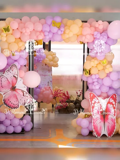 An entrance arch for a butterfly-themed party, made with pastel peach and purple balloons and accented with large pink butterfly cutouts.