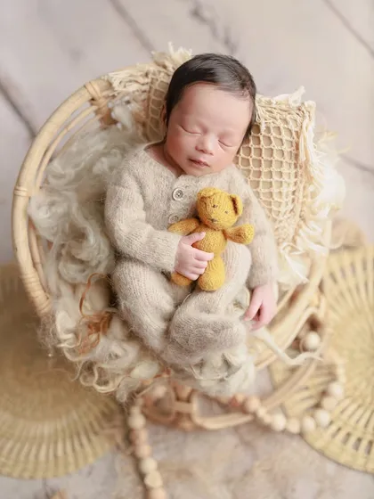 A cozy and rustic setup with neutral tones. This baby is dressed in a full knit romper, cuddling a teddy bear in a beautifully textured basket.