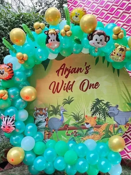 A 'Wild One' first birthday party with a jungle theme. This setup uses a custom printed backdrop and a balloon arch with green and gold chrome balloons and animal character foils.