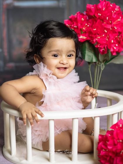 A happy baby girl in a pink ruffled romper sits in a white prop, surrounded by vibrant pink bougainvillea flowers.