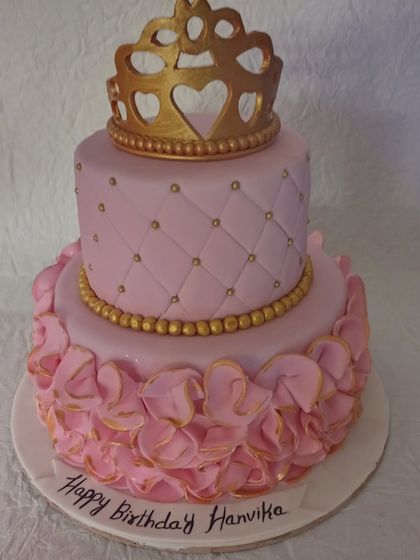 An elegant two-tiered princess cake in pink and gold. The top tier has a quilted pattern, while the bottom is covered in delicate ruffles, all topped with a golden crown.