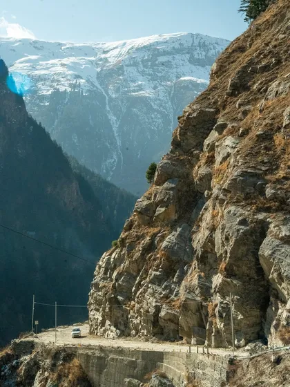 A lone white car navigates a narrow, treacherous road cut into the side of a massive cliff, with snow-capped mountains in the background. This shot powerfully conveys the scale and challenge of high-altitude driving.