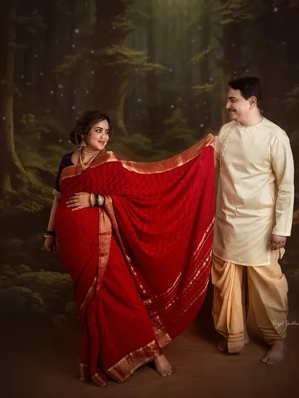 A playful, romantic moment between a couple, set against a mystical forest backdrop. The vibrant red saree stands out, creating a beautiful contrast that highlights the mother-to-be.