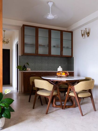The 5-seater circular dining table, creating a warm and inviting atmosphere for intimate meals. The olive-green fabric and vintage wall light complement the earthy tones of the living area.