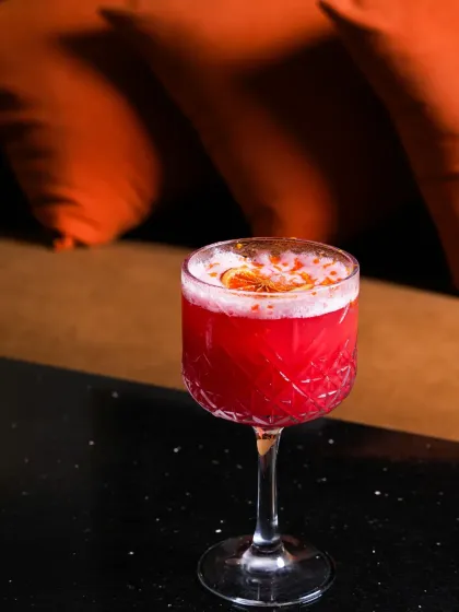 A single, vibrant red cocktail in an elegant glass, set against a dark, moody background with warm orange tones. This minimalist shot puts all the focus on the drink's color and classic presentation.