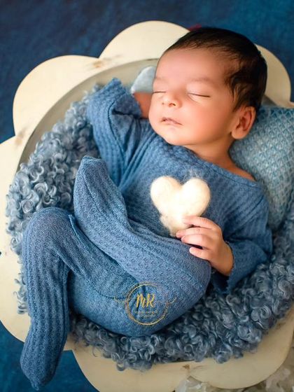 A newborn in a blue outfit holds a small white heart prop while sleeping in a flower-shaped bowl.