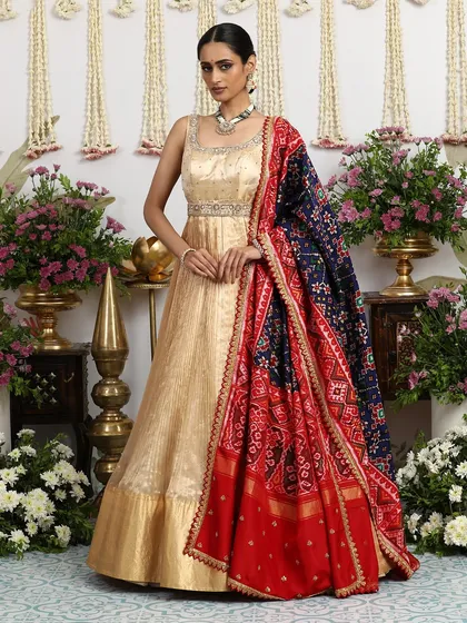 A floor-length Anarkali-style gown in gold silk, paired with a magnificent red and navy Bandhani dupatta. This is fusion wear at its most regal.