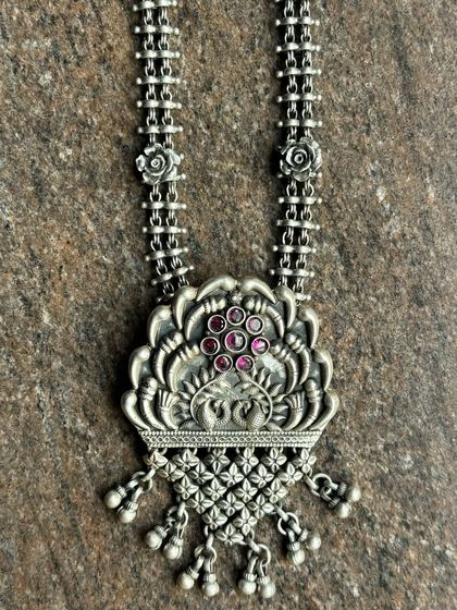 An oxidised silver pendant with a peacock-like floral design. The piece is studded with small pink stones, adding a subtle touch of colour.