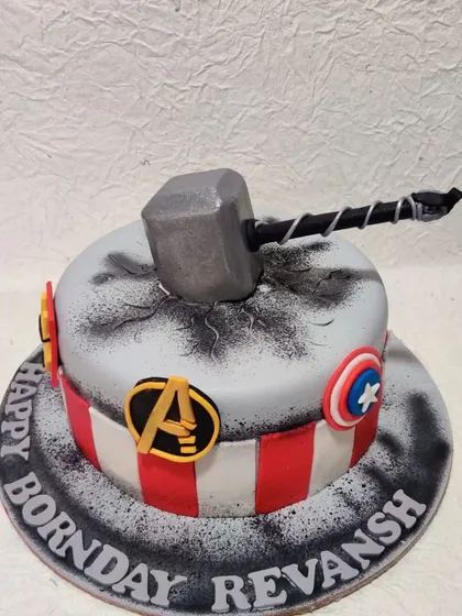 An Avengers-themed cake for a superhero fan. It features Thor's hammer crashing into the cake, along with the logos of Captain America and the Avengers.