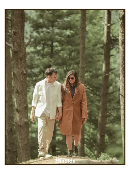 The couple walks hand-in-hand through a pine forest, sharing a look. A classic, candid walking shot that feels natural and full of connection.