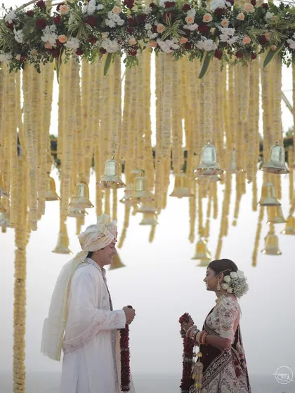 A serene varmala moment under a stunning floral chandelier adorned with traditional bells. The couple shares a look of love before exchanging garlands.