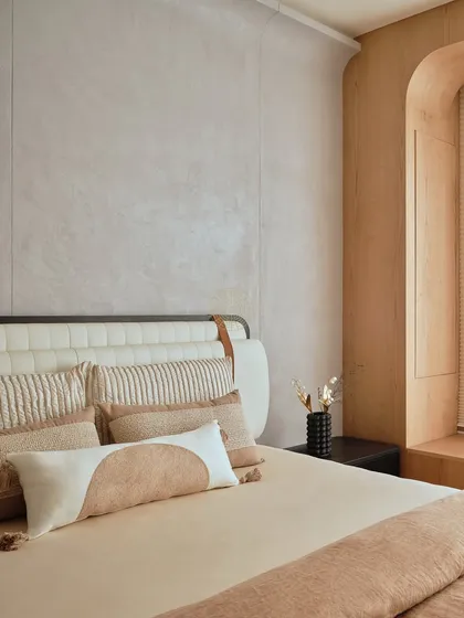 A cozy bedroom in a Bangalore home featuring a textured wall, a custom upholstered headboard with leather strap details, and a warm, earthy color palette. The arched window nook adds architectural interest.