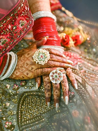A detailed shot of a bride's hands, showcasing her elaborate rings and henna against the rich texture of her lehenga.