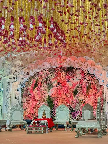 A mandap designed to be a blooming masterpiece. The backdrop is a dense wall of intricate florals, complemented by regal silver seating and a ceiling of hanging flower strings. 🌸🩷