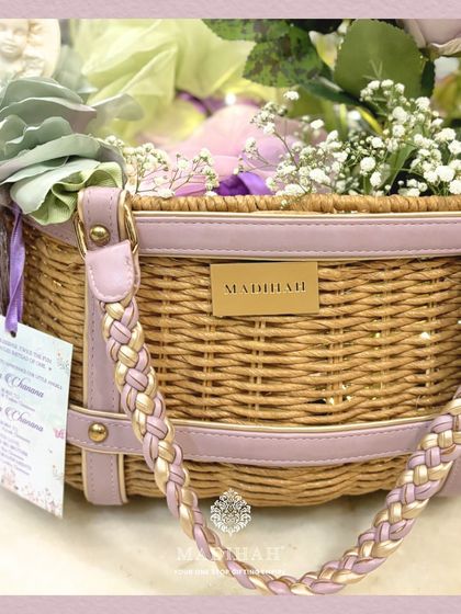A detailed close-up of the Cane Arrival Basket, focusing on the personalized announcement card and the braided handle. The combination of natural cane with lavender leather and gold accents adds a touch of modern elegance.