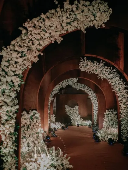 A side angle of the Sangeet entrance tunnel, highlighting the contrast between the rustic arches and the delicate white floral arrangements. This is an example of our custom fabrication capabilities.