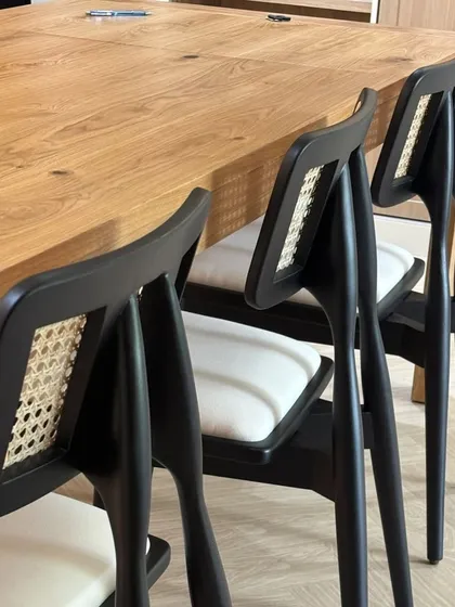 A close-up of my Jeanneret-style dining chairs with a black finish and white seat cushions, paired with a solid oak table. The caned backrest provides flexible comfort.