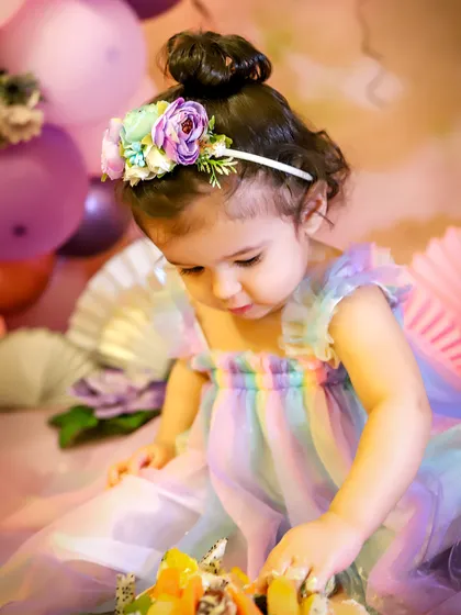 A close-up of the birthday girl curiously touching her fruit-topped cake. The details, from the floral headband to the rainbow dress, are all part of the fun.