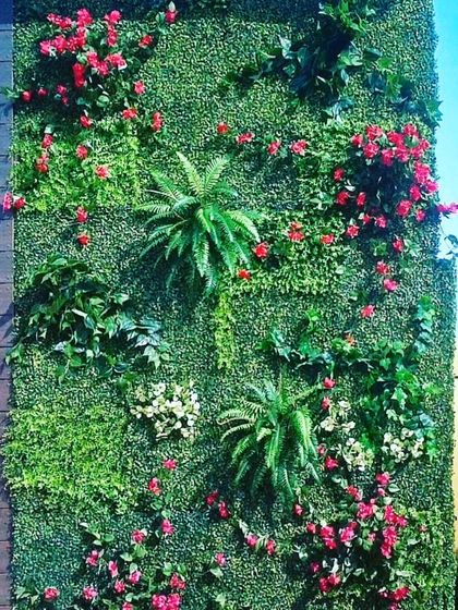 An outdoor green wall featuring a mix of ferns, broad leaves, and clusters of small red artificial flowers. This adds color and variety to a standard vertical garden installation.