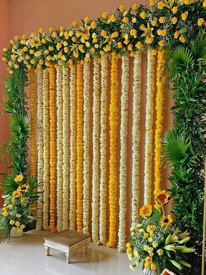 A classic backdrop for a haldi ceremony or pooja, featuring an arch of yellow roses and hanging strings of marigold and tuberose flowers.