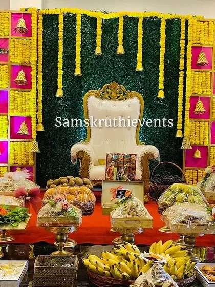 A vibrant baby shower backdrop in yellow and pink, with hanging marigold strings and traditional bells. The foreground shows a lavish display of covered offering trays, ready for the rituals.