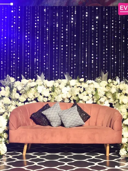 A chic seating area for a Sangeet, with a blush-colored sofa set against a backdrop of hanging silver sequins and framed by elegant white floral arrangements.