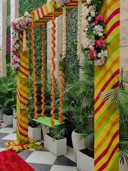 A detailed look at a decorated jhoola (swing) for a Teej festival. The swing is adorned with marigold garlands and set against a colorful striped pillar and lush greenery.