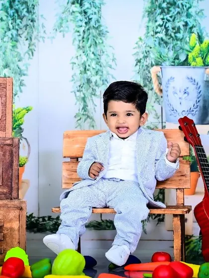 A one-year-old boy in a suit, sitting on a bench in a garden-themed setup.
