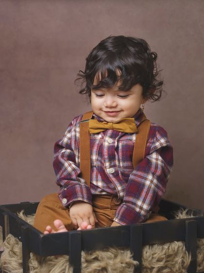 A quiet, gentle moment captured in the studio. This little boy, dressed in a classic plaid shirt and bow tie, looks down with a sweet expression, showcasing a different side of his personality.