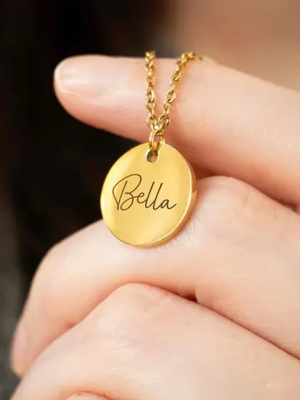 A hand holding a gold round pendant engraved with the name 'Bella' in a beautiful script. This highlights the delicate and personal nature of the engraving.