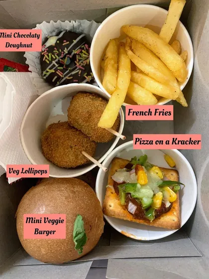 A close-up of a perfectly portioned snack box for a kid's party. It includes a mini veggie burger, veg lollipops, french fries, pizza on a cracker, and a mini chocolate doughnut, a complete and happy meal for any child.