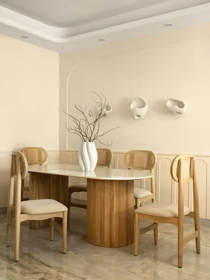 The dining area of Casa Levante embraces a Wabi-Sabi elegance. A fluted wooden table base is topped with marble, surrounded by cane-back chairs. The wall is adorned with minimalist ceramic art.