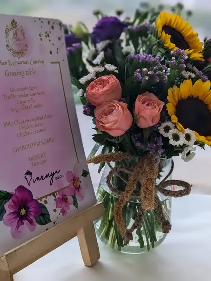 The custom menu for the Varnya salon launch, surrounded by a beautiful bouquet of sunflowers and roses. The menu details the gourmet tacos, sliders, and charcuterie board we prepared.