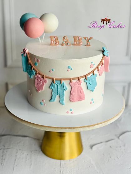 A classic baby shower cake with a clothesline of tiny fondant outfits. The pastel pink and blue dots and balloon toppers add to the sweet and celebratory feel.