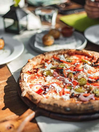 A beautiful pizza served on a wooden board, basking in the sunlight. It's topped with jalapeños, red peppers, and feta cheese for a vibrant and flavorful combination.
