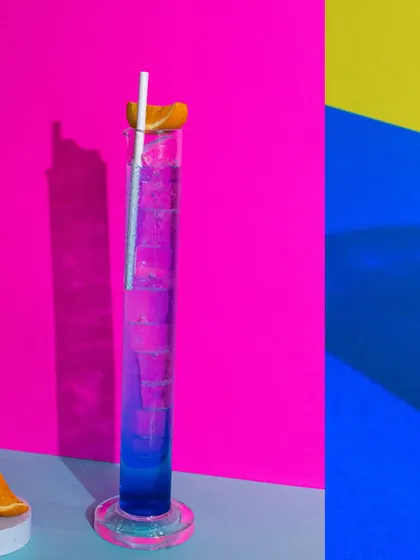 A tall, vibrant blue and purple cocktail in a science beaker-style glass. The bold, blocky background colours make this a really fun and unconventional drink shot.