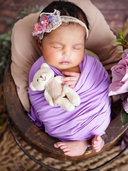 A peaceful newborn wrapped in a purple swaddle, holding a tiny teddy bear close while sleeping in a rustic basket.